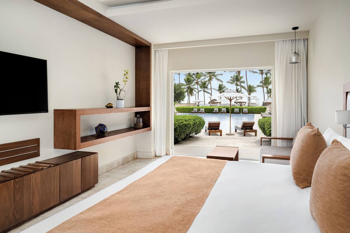 A modern hotel room with a large bed, a TV mounted on the wall, a wooden console table, and a view of the pool and palm trees outside.