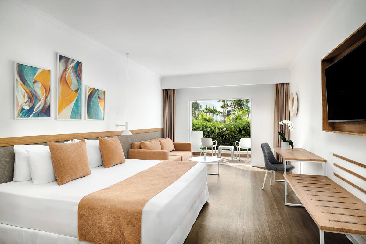A modern hotel room with a large bed, a desk, and a view of greenery outside.