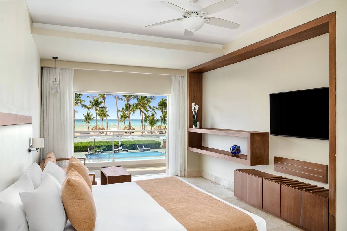 A hotel room with a bed, a TV, a wooden cabinet, a small table, and a view of the beach and pool.