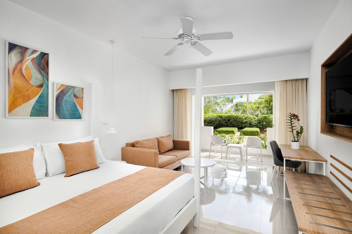 A modern hotel room with a bed, a sofa, a desk, and a balcony with outdoor furniture.