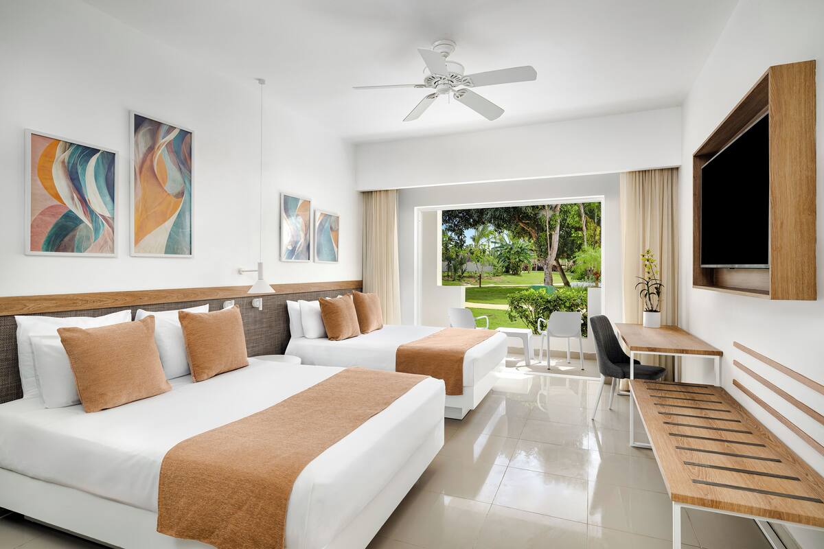 A hotel room with two beds, a desk, and a TV. There are abstract paintings on the wall and a view of greenery outside.