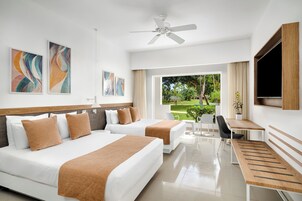 Quarto de hotel com duas camas, uma escrivaninha e uma TV. Há quadros abstratos na parede e vista para vegetação lá fora.