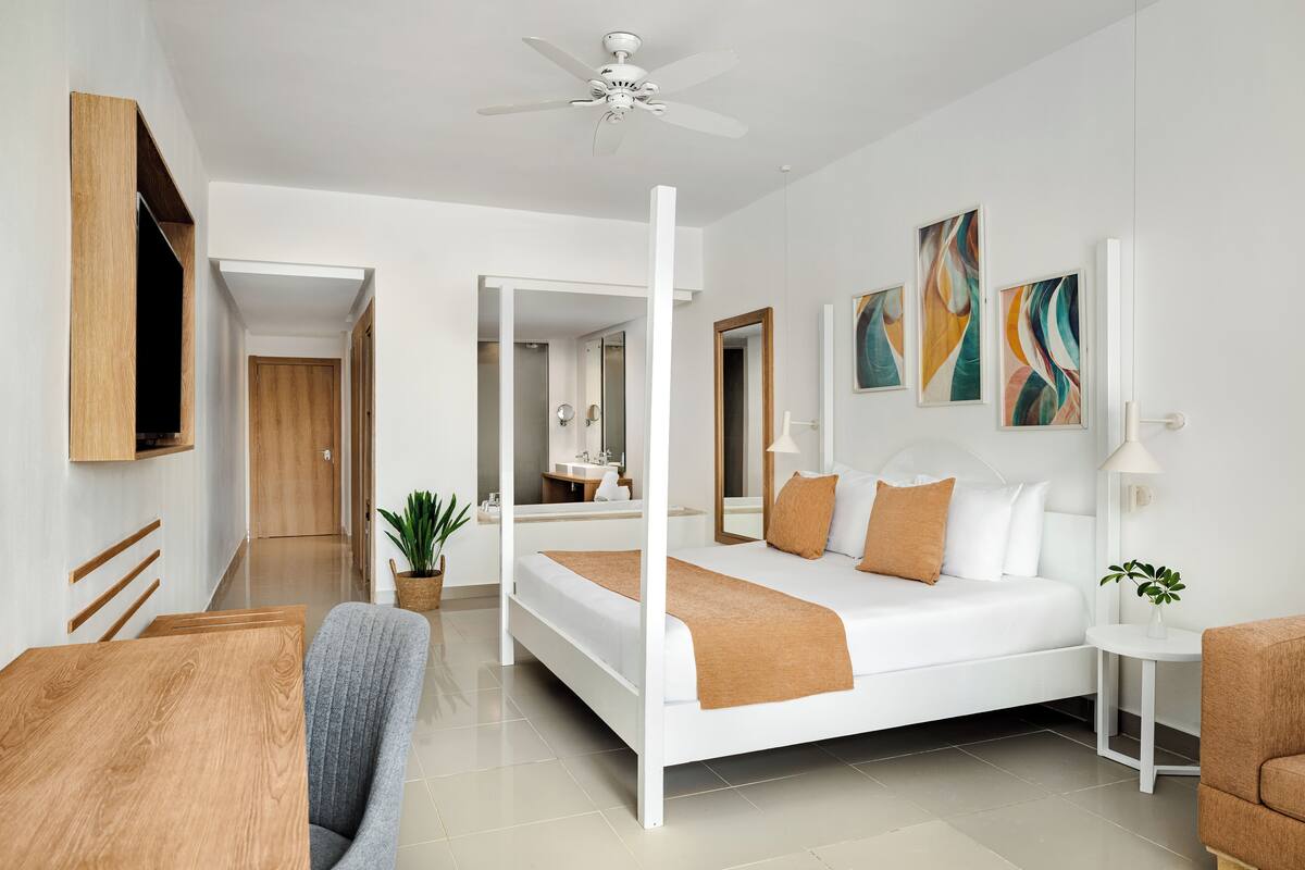 A modern hotel room with a large bed, a ceiling fan, a small table, a chair, and a potted plant.
