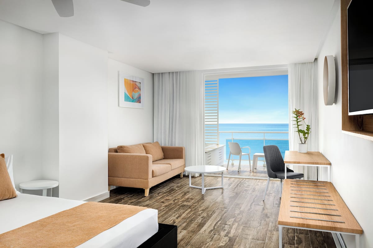 A modern hotel room with a bed, a sofa, a small table, a chair, and a view of the ocean.