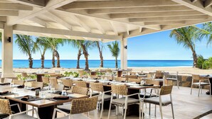 An outdoor dining area with tables and chairs, overlooking a beach with palm trees and the ocean.