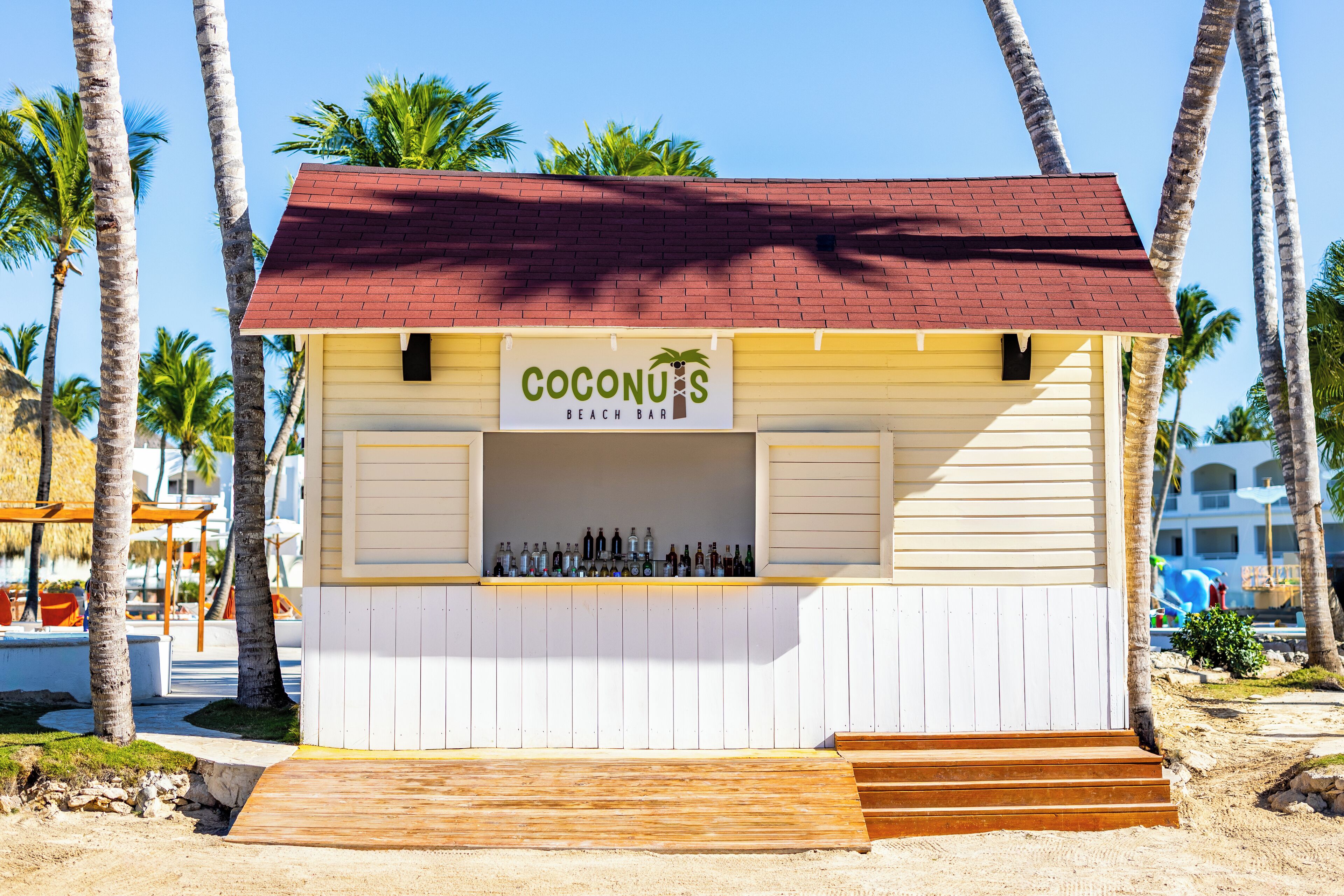 Un bar sulla spiaggia chiamato 'Coconuts', con un tetto rosso, scalini in legno e una vetrina piena di bottiglie.