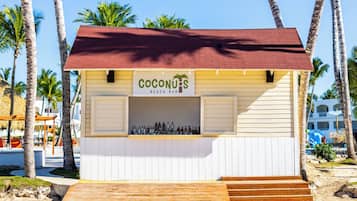 Un bar sulla spiaggia chiamato 'Coconuts', con un tetto rosso, scalini in legno e una vetrina piena di bottiglie.