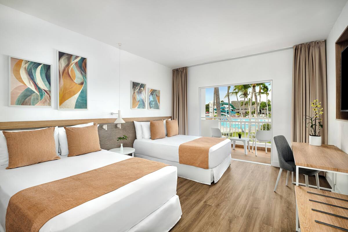 A hotel room with two beds, a dining table, and a view of a pool and palm trees.