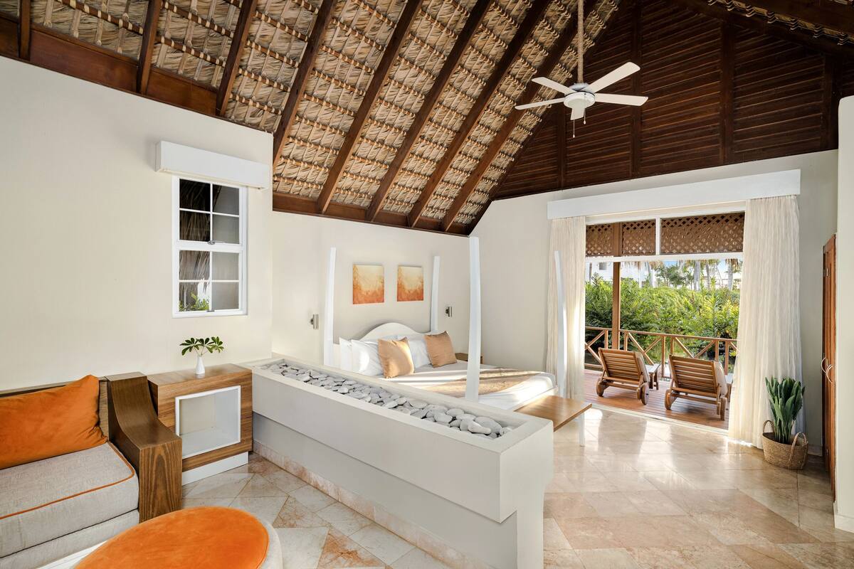 A spacious bedroom with a large bed, a sitting area with a sofa and ottoman, a balcony with wooden chairs, and a ceiling fan.