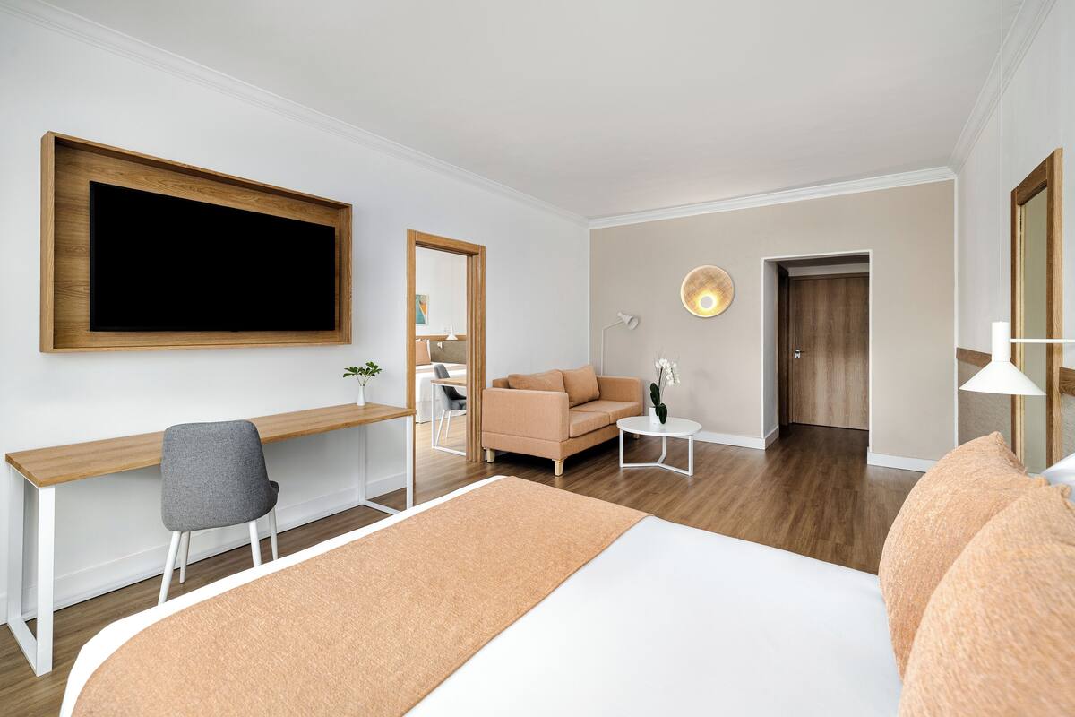 A modern hotel room with a large bed, a desk, a sofa, and a TV mounted on the wall.