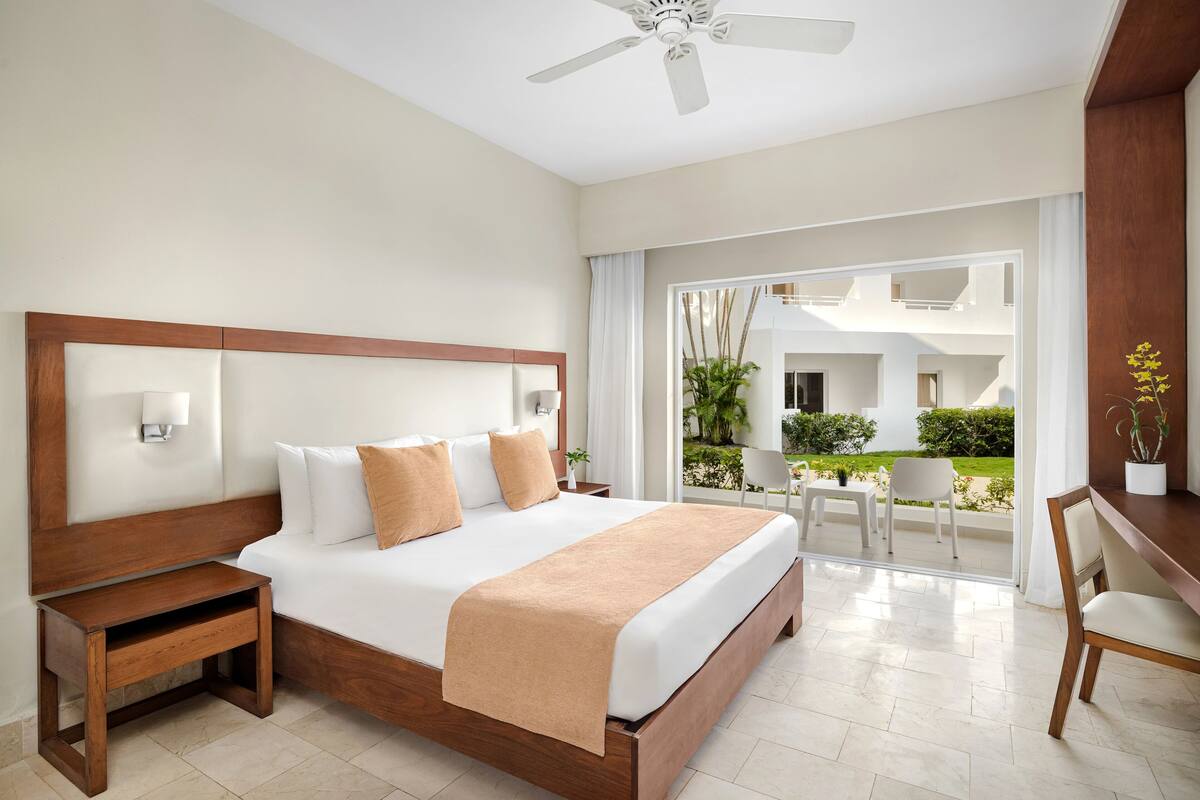 A hotel room with a large bed, a wooden nightstand, a desk with a chair, and a view of the outdoor area.