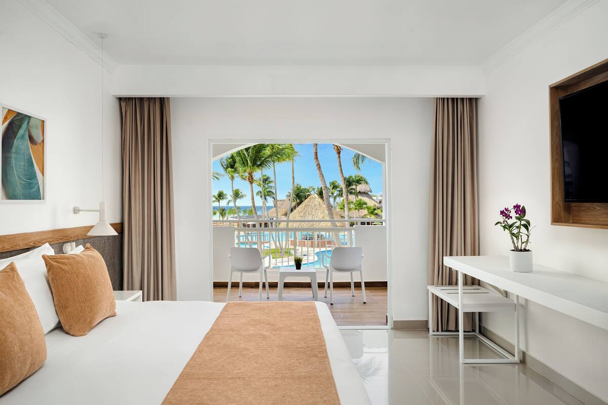 A hotel room with a bed, a desk, and a view of palm trees and a pool.