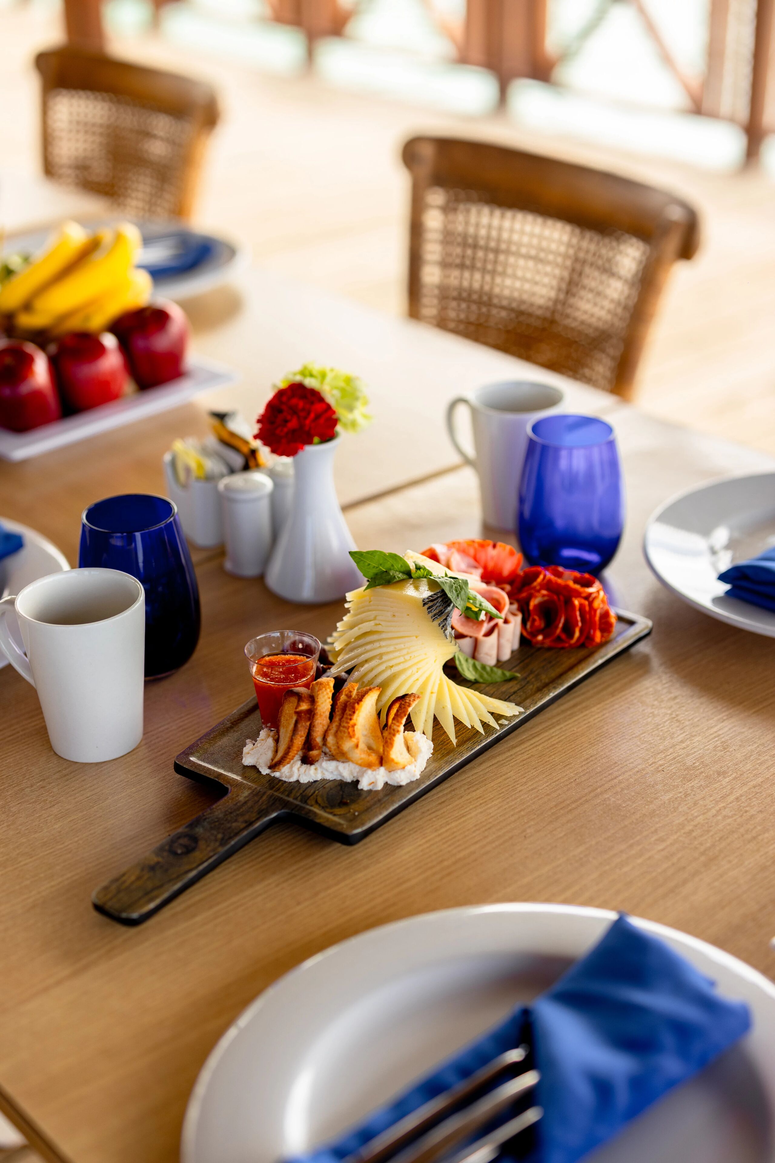 Une table à manger sur laquelle on trouve une variété d’aliments, notamment un plateau de fromages, des fruits et un petit vase avec des fleurs.