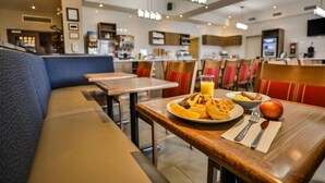 Une salle à manger d’hôtel avec des tables et des chaises, une table préparée pour le petit-déjeuner et une cuisine en arrière-plan.