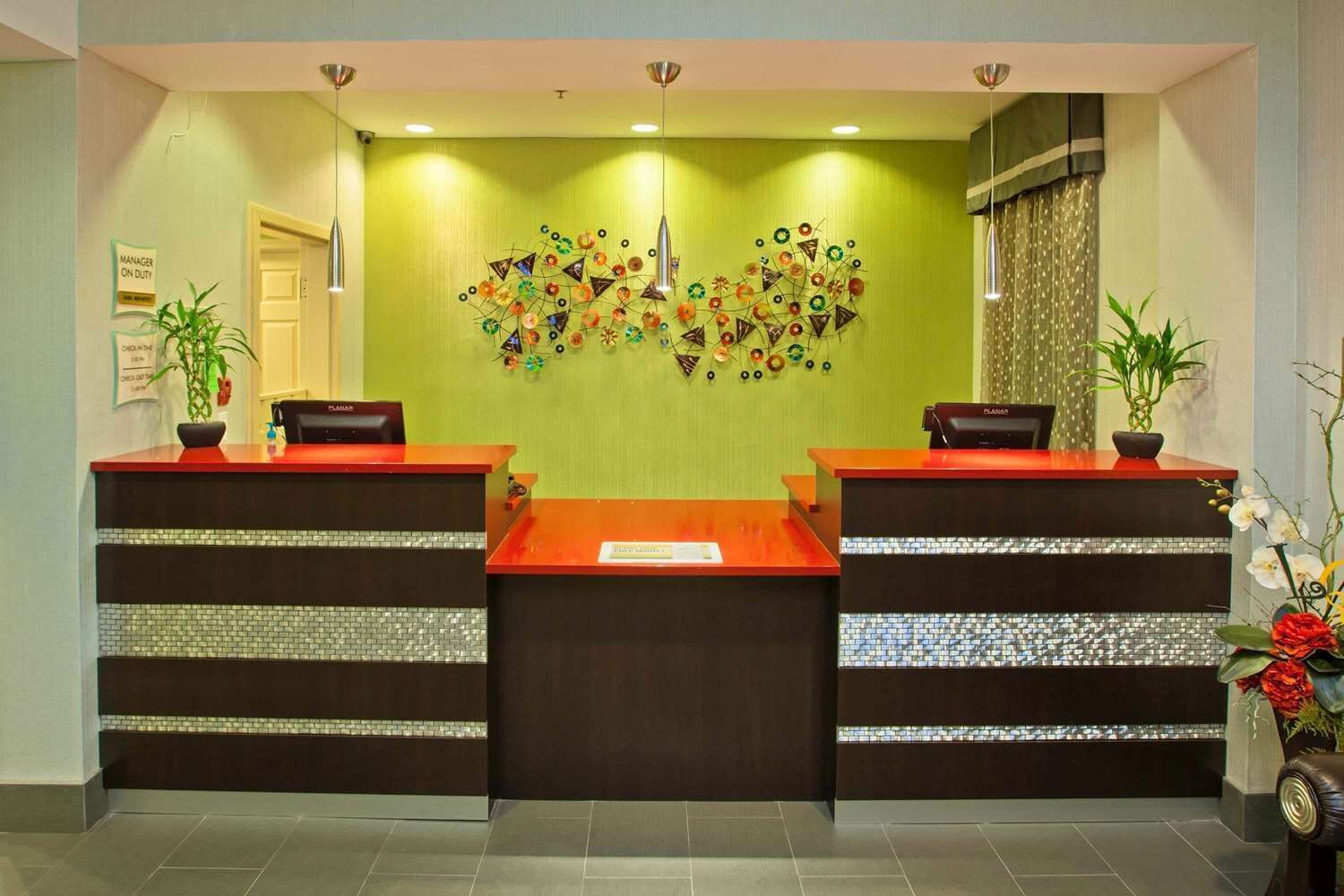 Zona de recepción con dos mostradores, una pared verde con una obra de arte metálica decorativa y plantas en macetas.