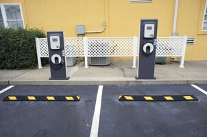 Duas estações de carregamento para veículos elétricos, com uma cerca de grade branca e um edifício amarelo ao fundo.