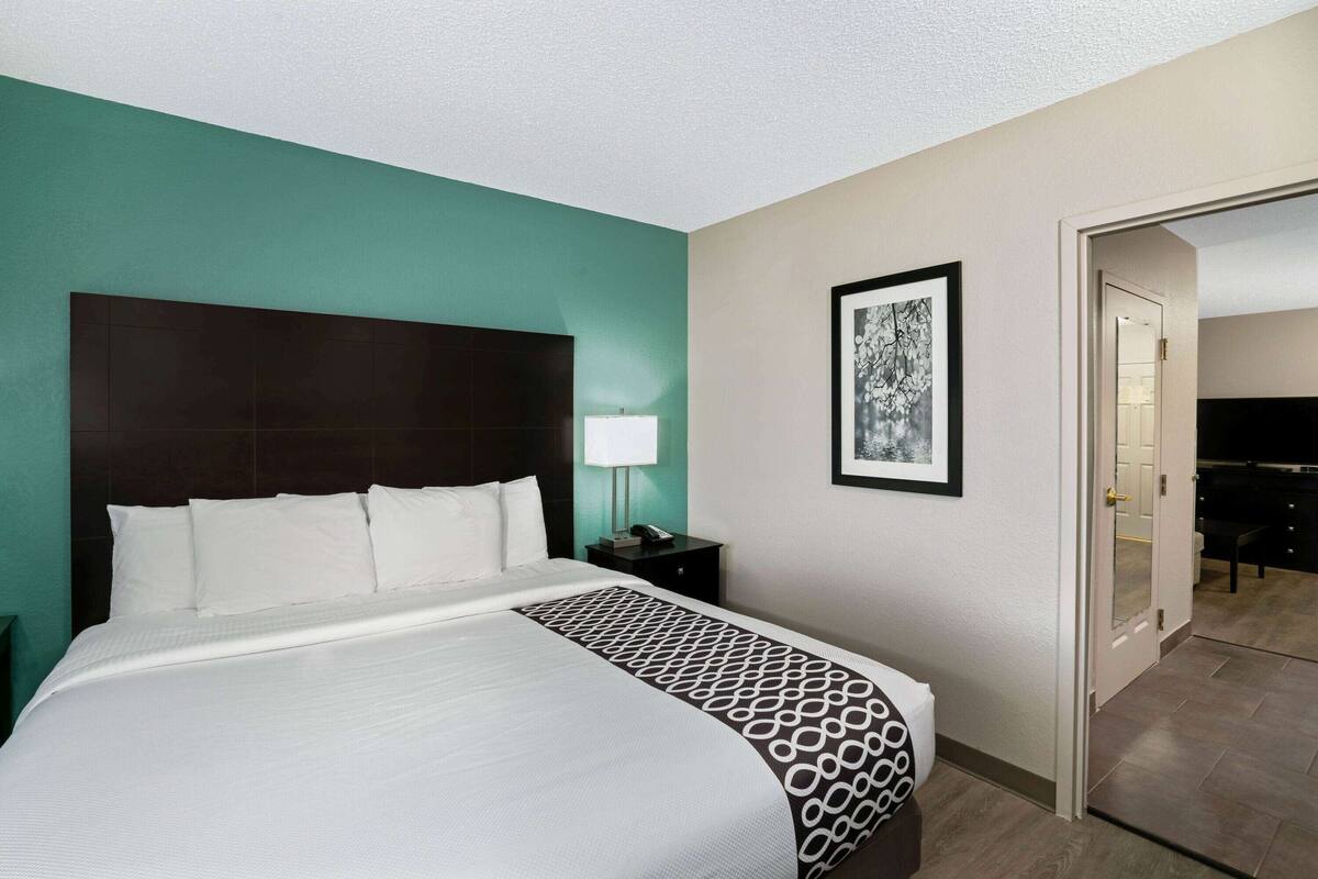 Une chambre d’hôtel avec un grand lit, une table de chevet avec une lampe, un tableau encadré au mur et une porte donnant sur une salle de bain.