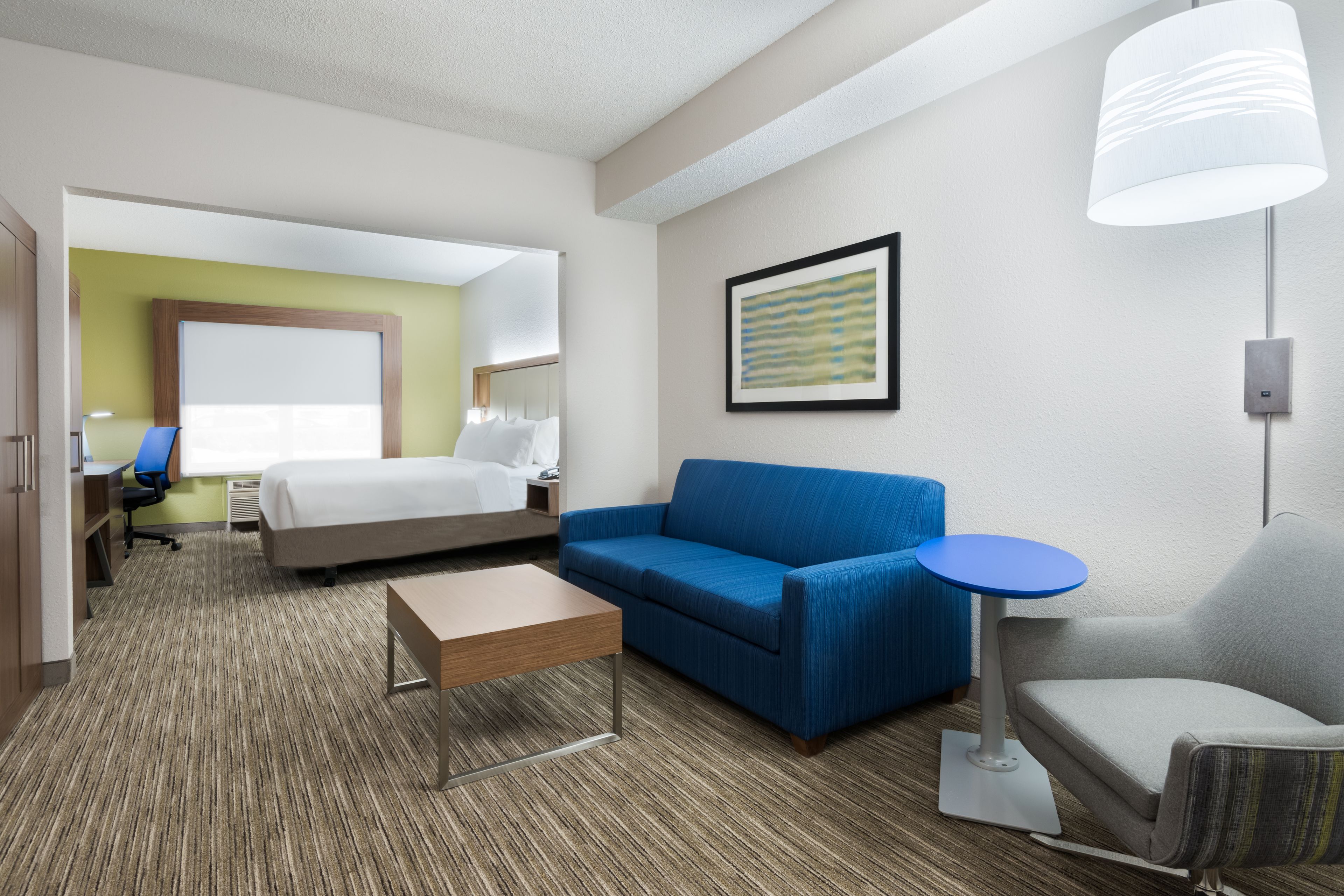 A hotel room with a bed, a blue sofa, a small table, a grey armchair, a blue side table, a lamp, and a framed picture on the wall.