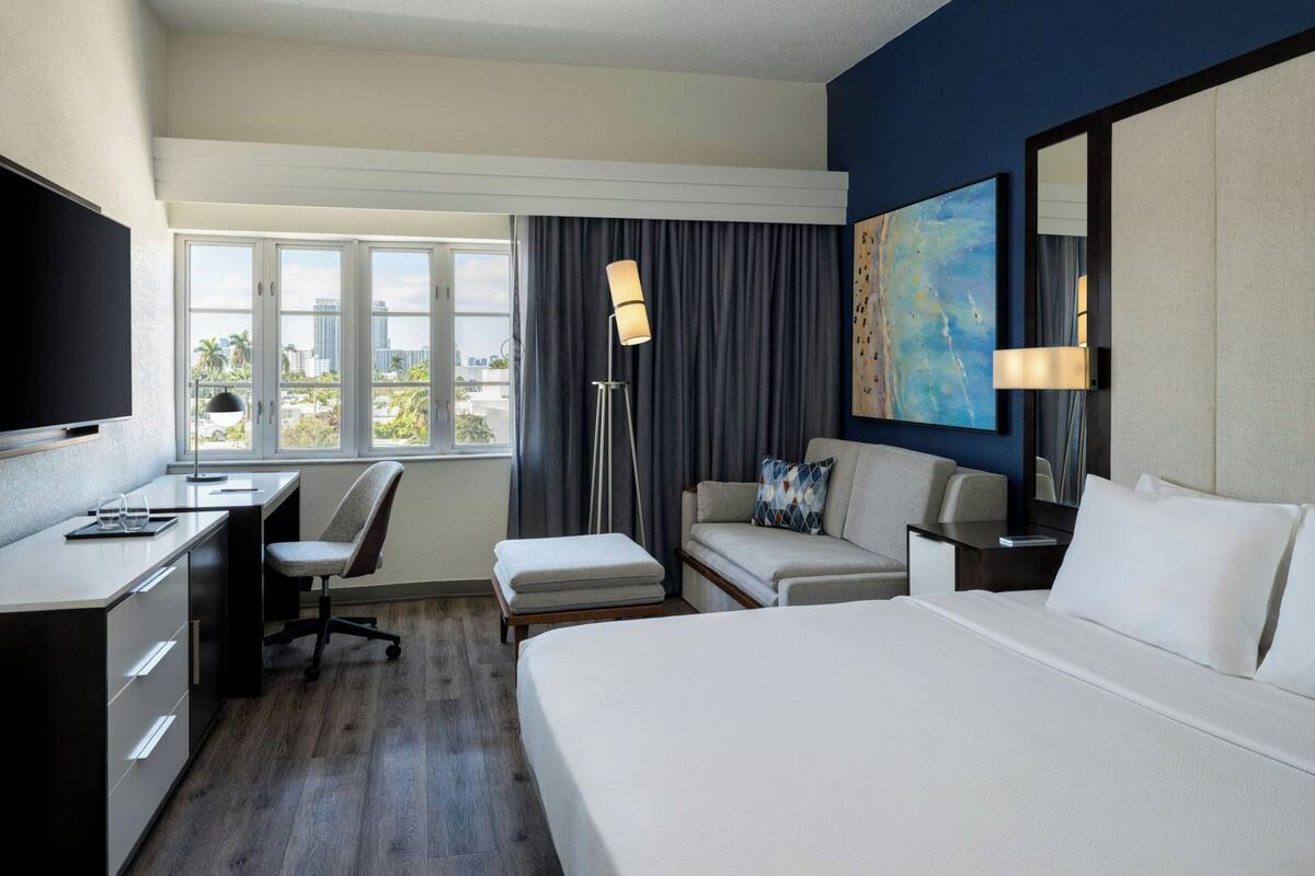 Habitación de hotel con cama, escritorio, silla y un ventanal que ofrece vistas a la ciudad.