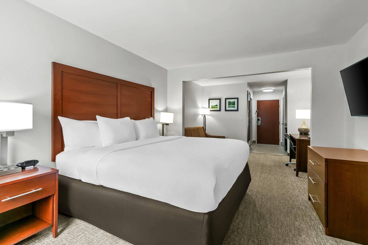 A hotel room with a large bed, a wooden headboard, a desk with a lamp, a dresser, and a TV mounted on the wall.