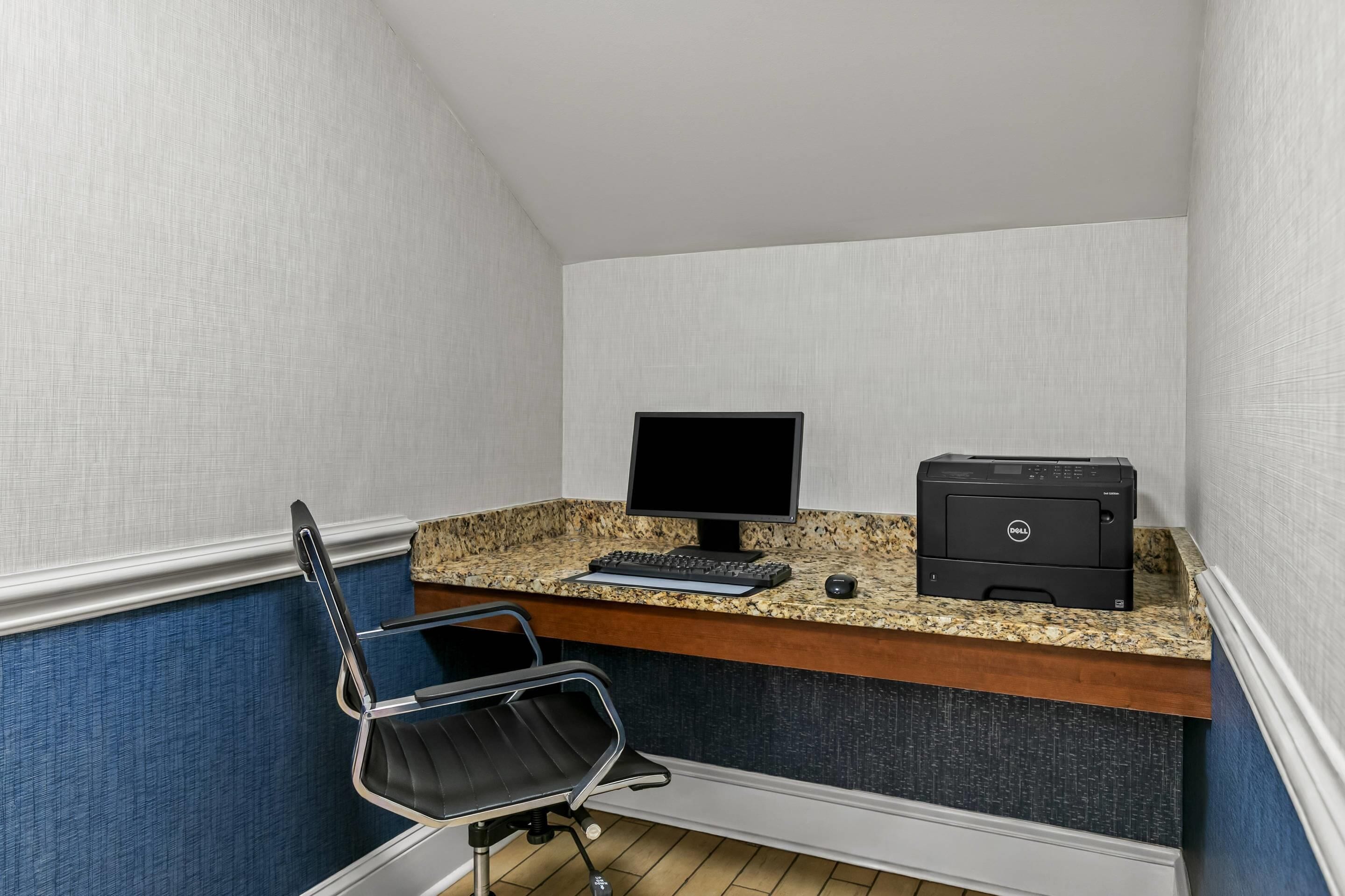 Een kleine kantoorruimte met een bureau, stoel, computerscherm, toetsenbord en printer.