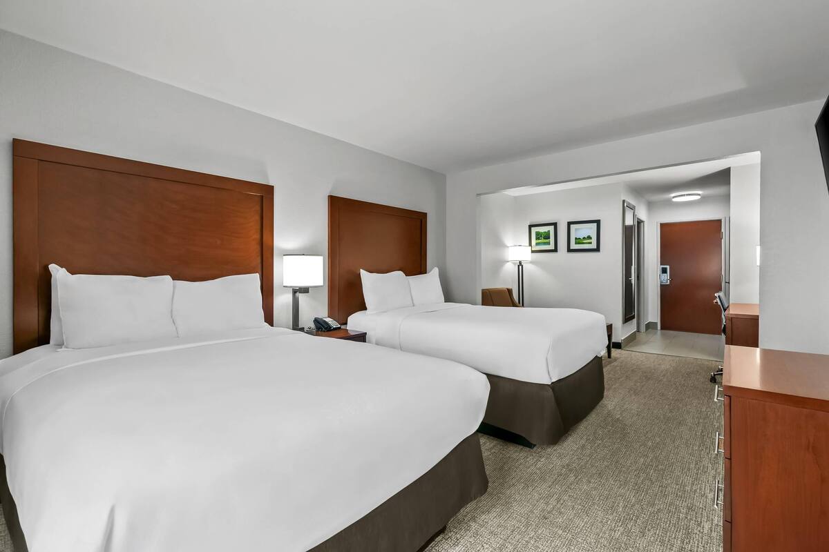 A hotel room with two beds, a desk, a chair, a lamp, and framed pictures on the wall.