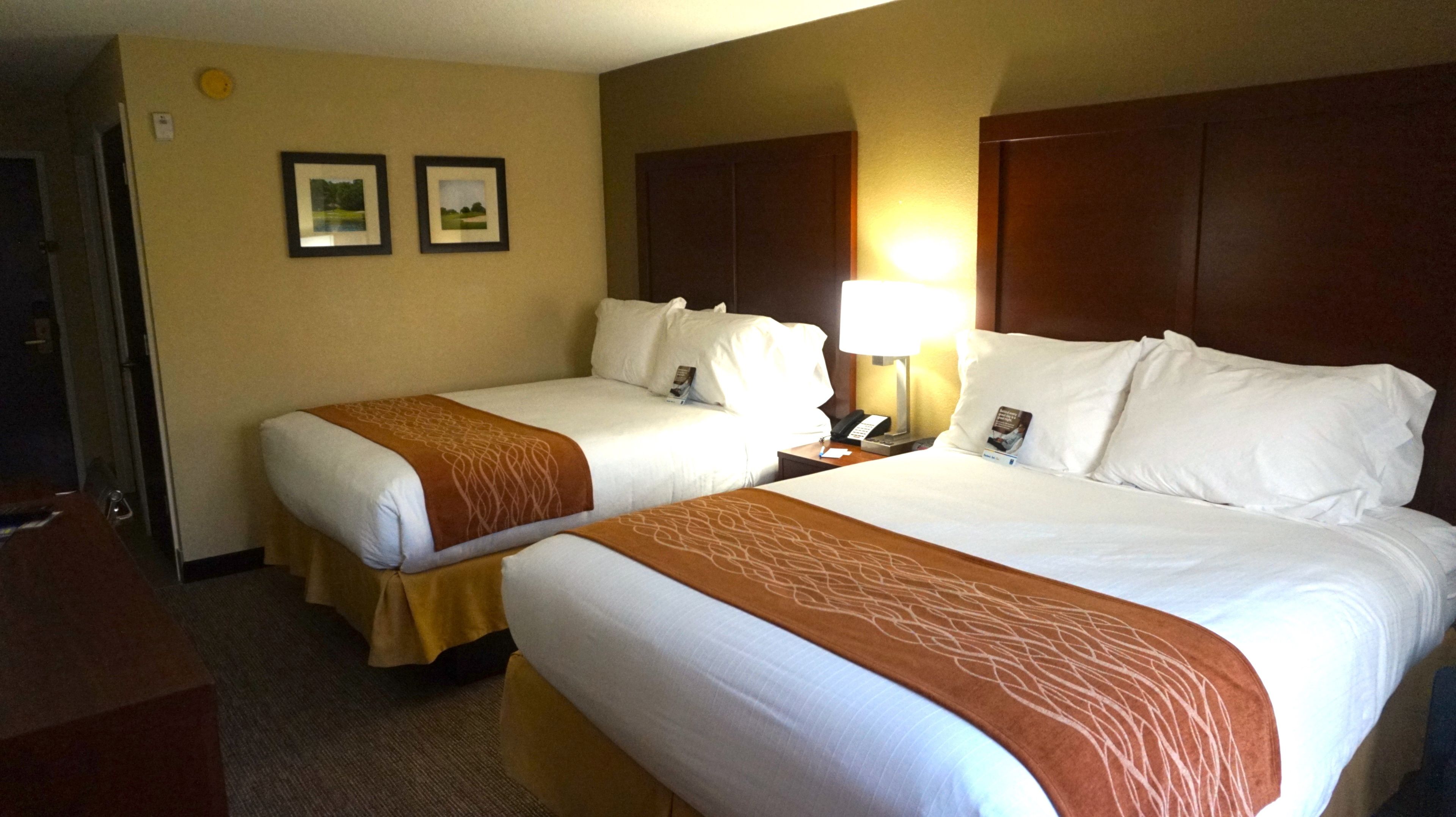 A hotel room with two beds, a bedside table with a lamp, and two framed pictures on the wall.