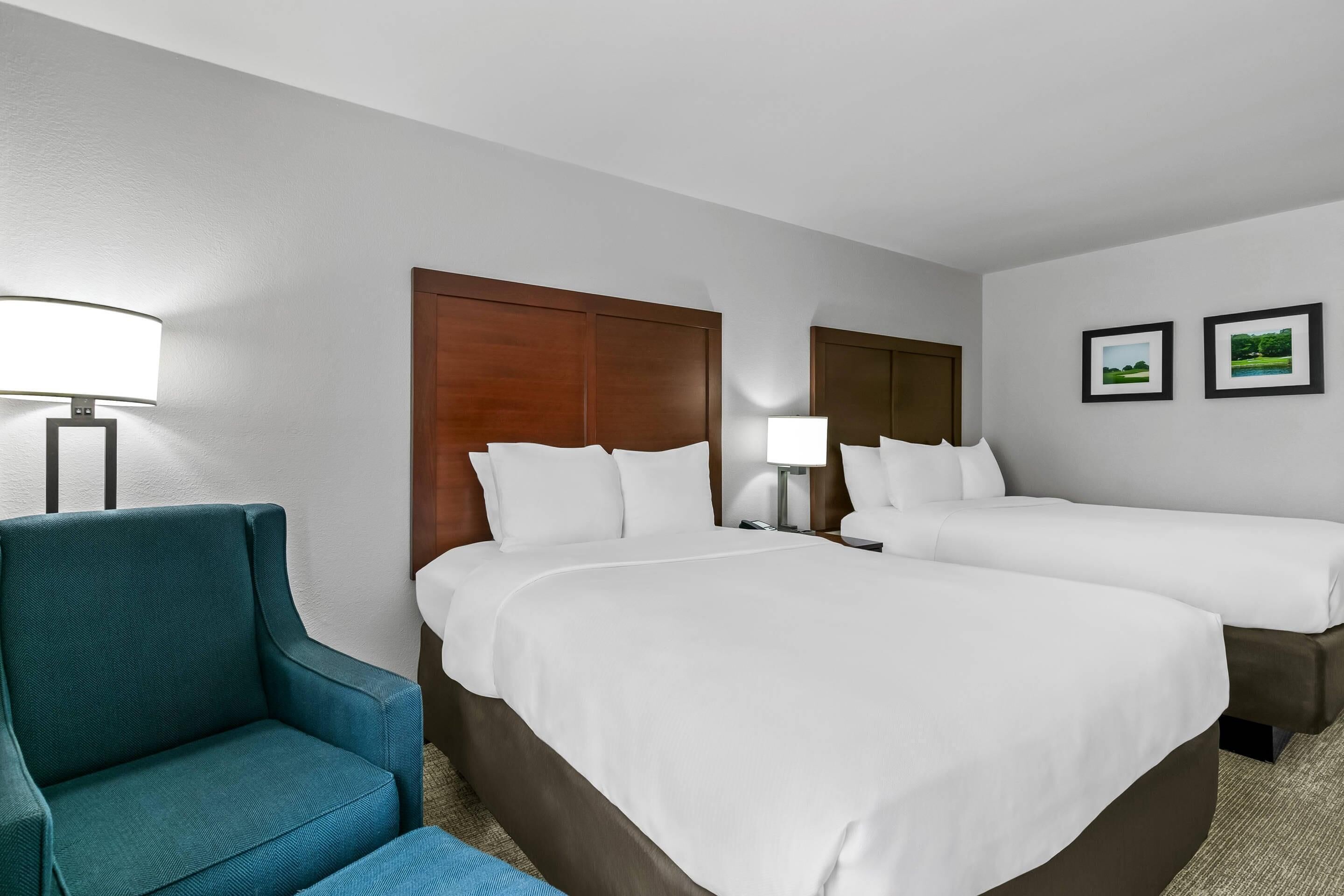 A hotel room with two beds, a blue armchair, a nightstand with a lamp, and two framed pictures on the wall.