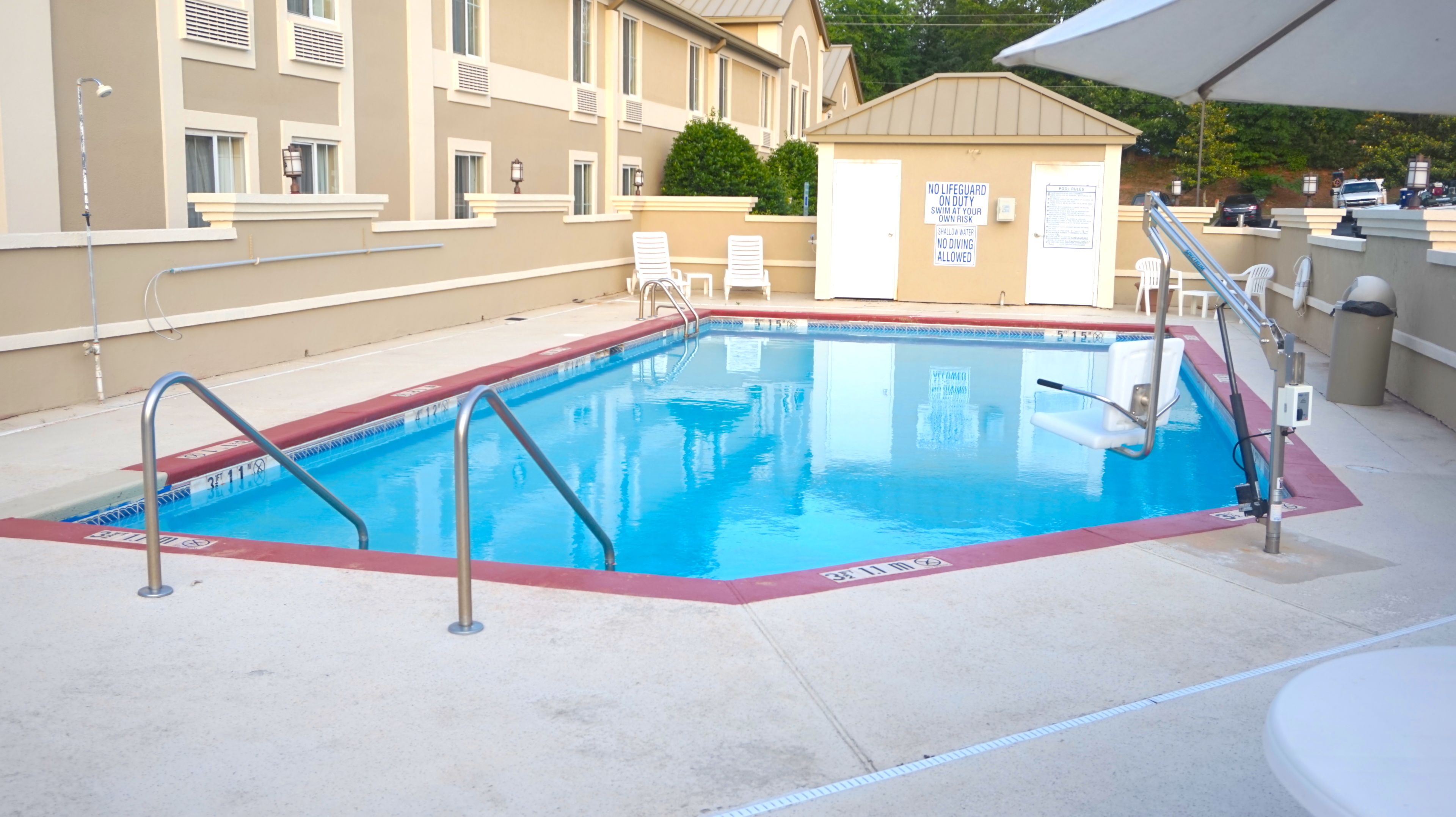 Una piscina rectangular con borde rojo, rodeada por una solana de concreto. Hay dos sillas de descanso blancas y un pequeño edificio con un cartel que indica que no hay salvavidas de servicio.