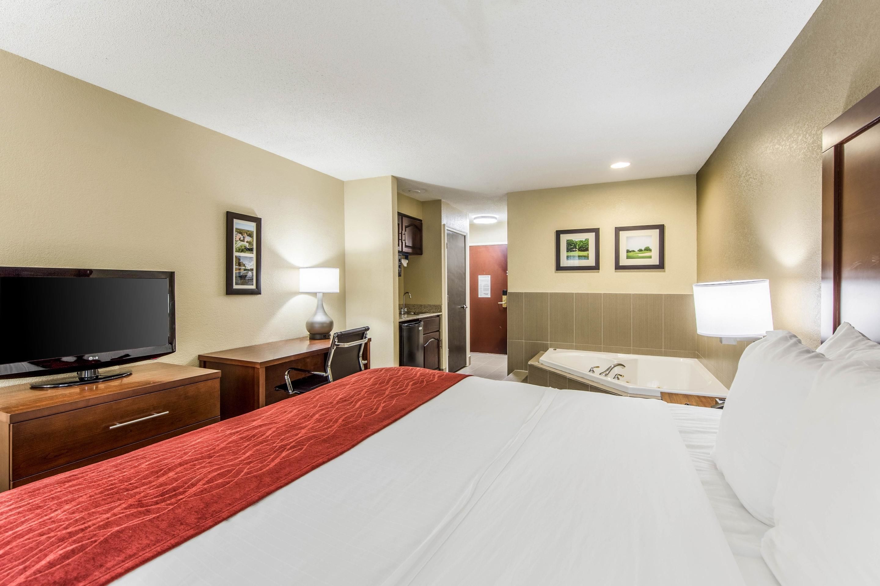 Quarto de hotel com cama, escrivaninha com televisão, banheiro com banheira e cozinha compacta.