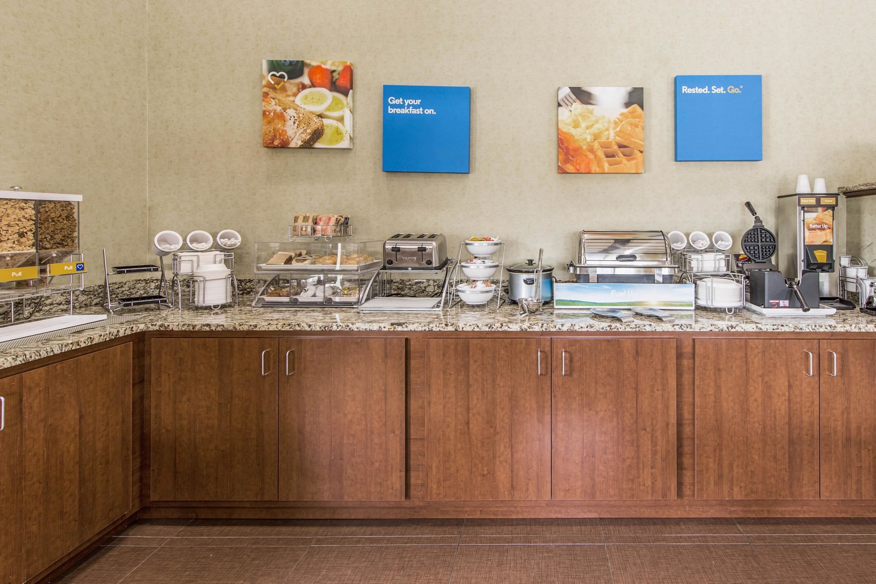 A breakfast area with a granite countertop, coffee machines, a waffle maker, and a bread display.