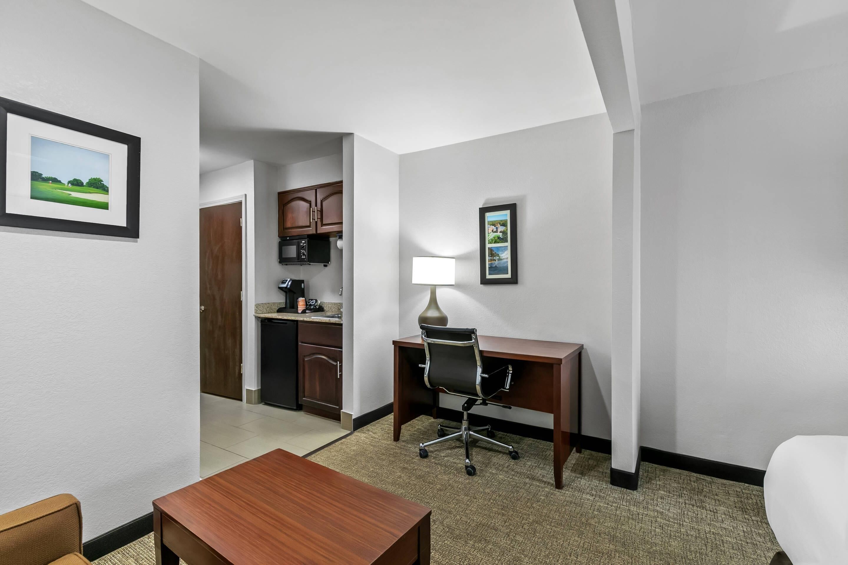 A hotel room with a desk, chair, lamp, and a view of the kitchen area.