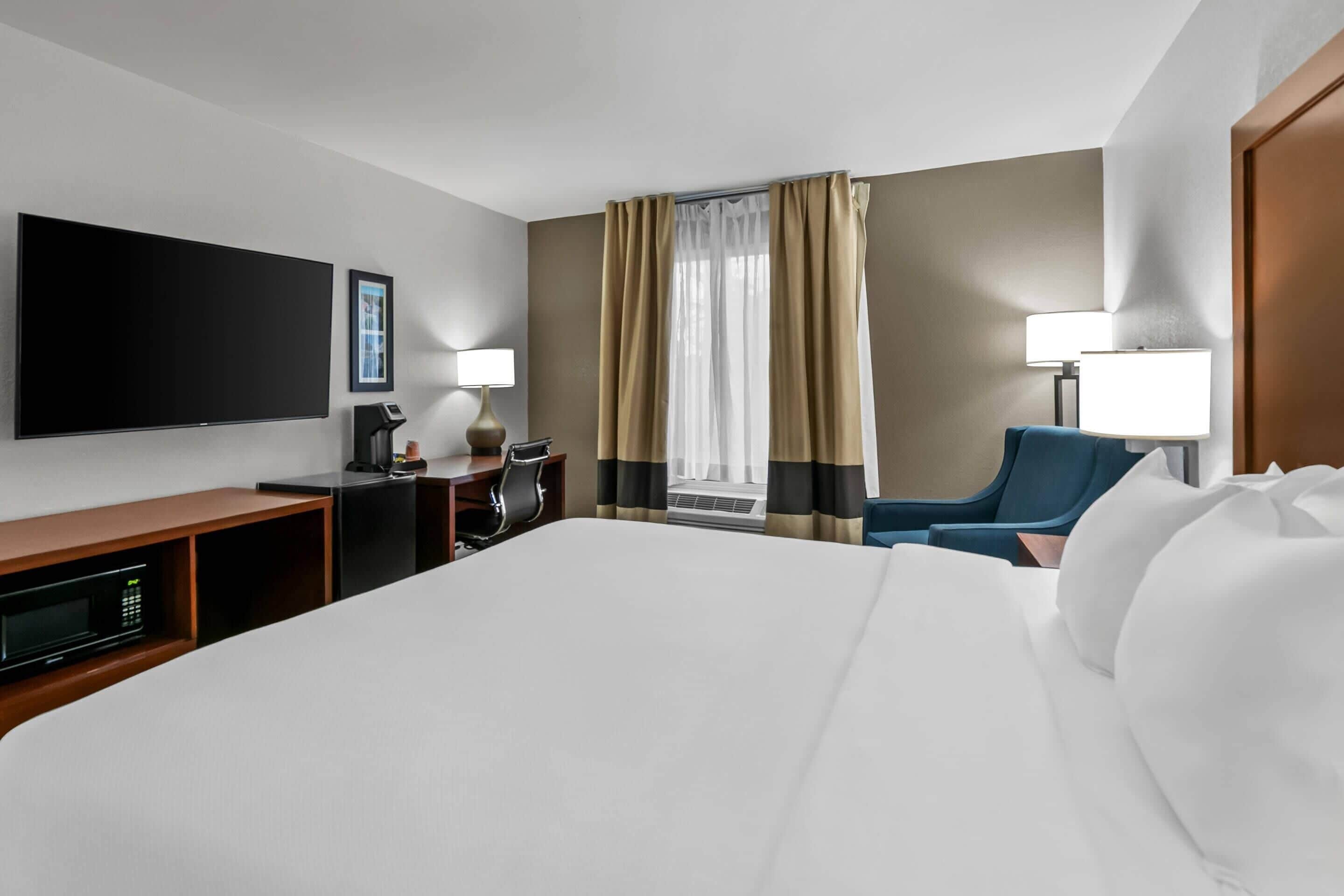 A hotel room with a large bed, a flat-screen TV, a desk with a lamp, a chair, and a window with curtains.