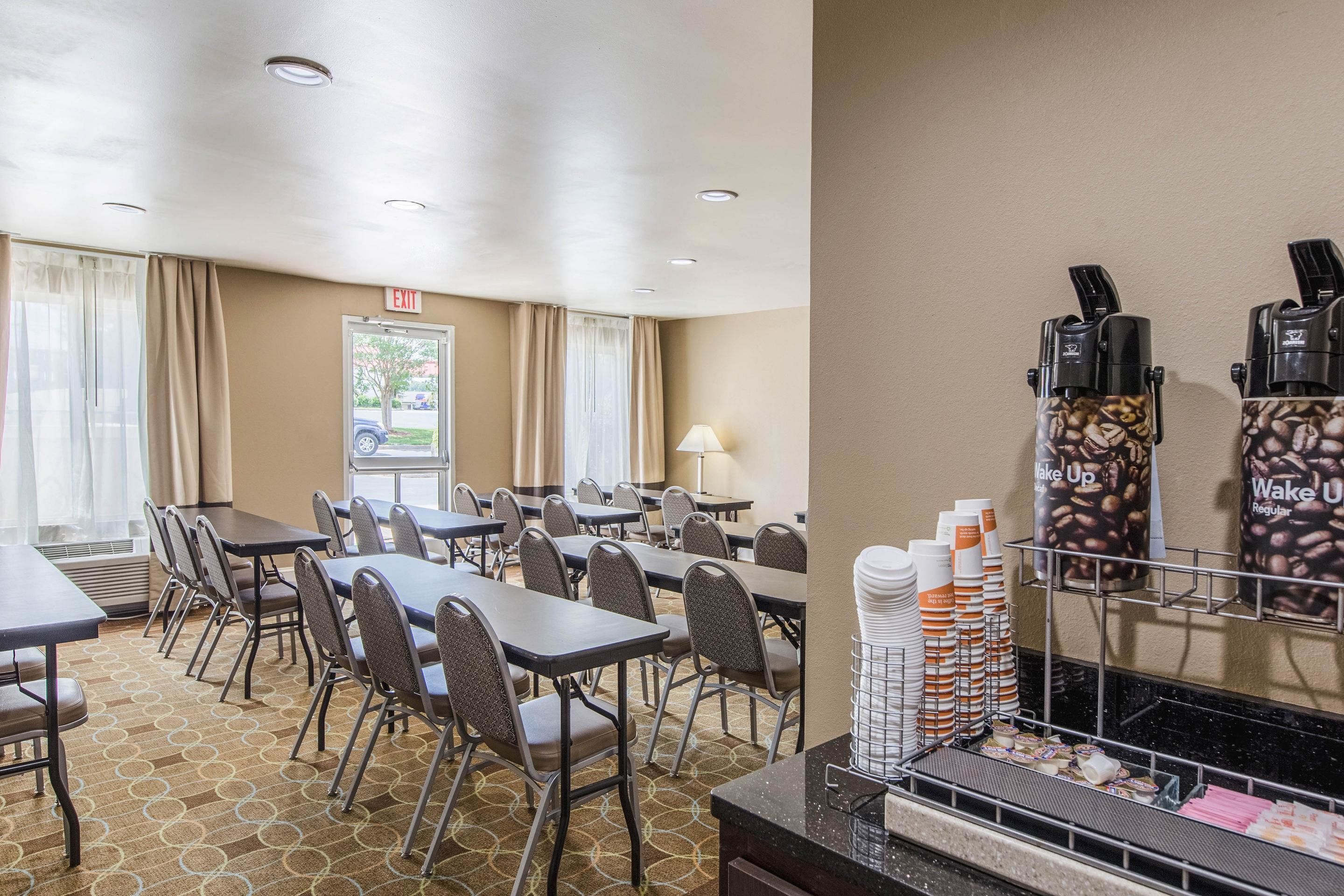 A hotel dining area with tables and chairs, an exit sign, and a coffee machine.
