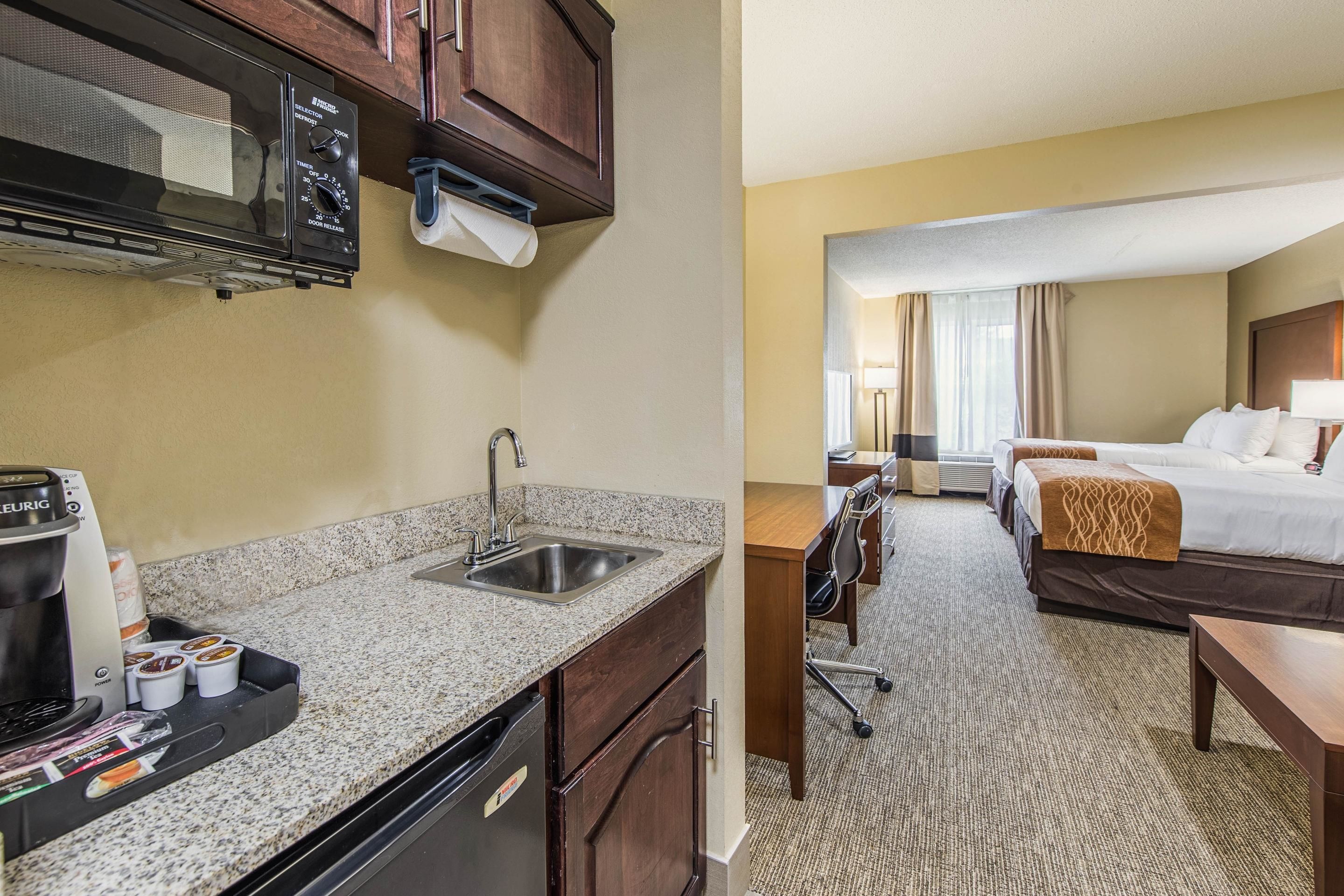A hotel room with a kitchenette, including a microwave, coffee maker, and sink. There is a bed with a headboard, a desk, and a chair. The room has a carpeted floor and a window with curtains.