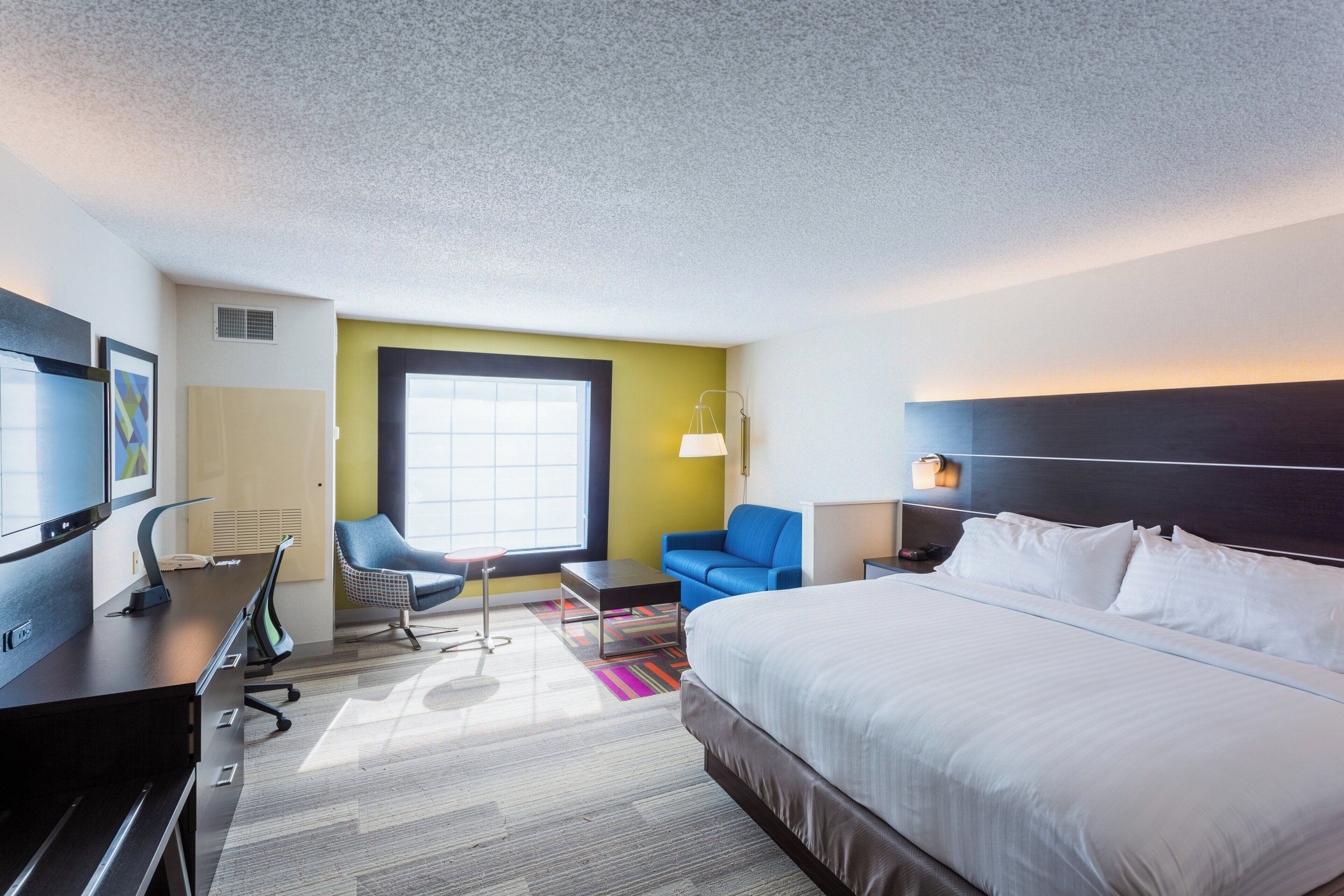 A hotel room with a bed, a desk with a computer, a chair, a small table, and a window.