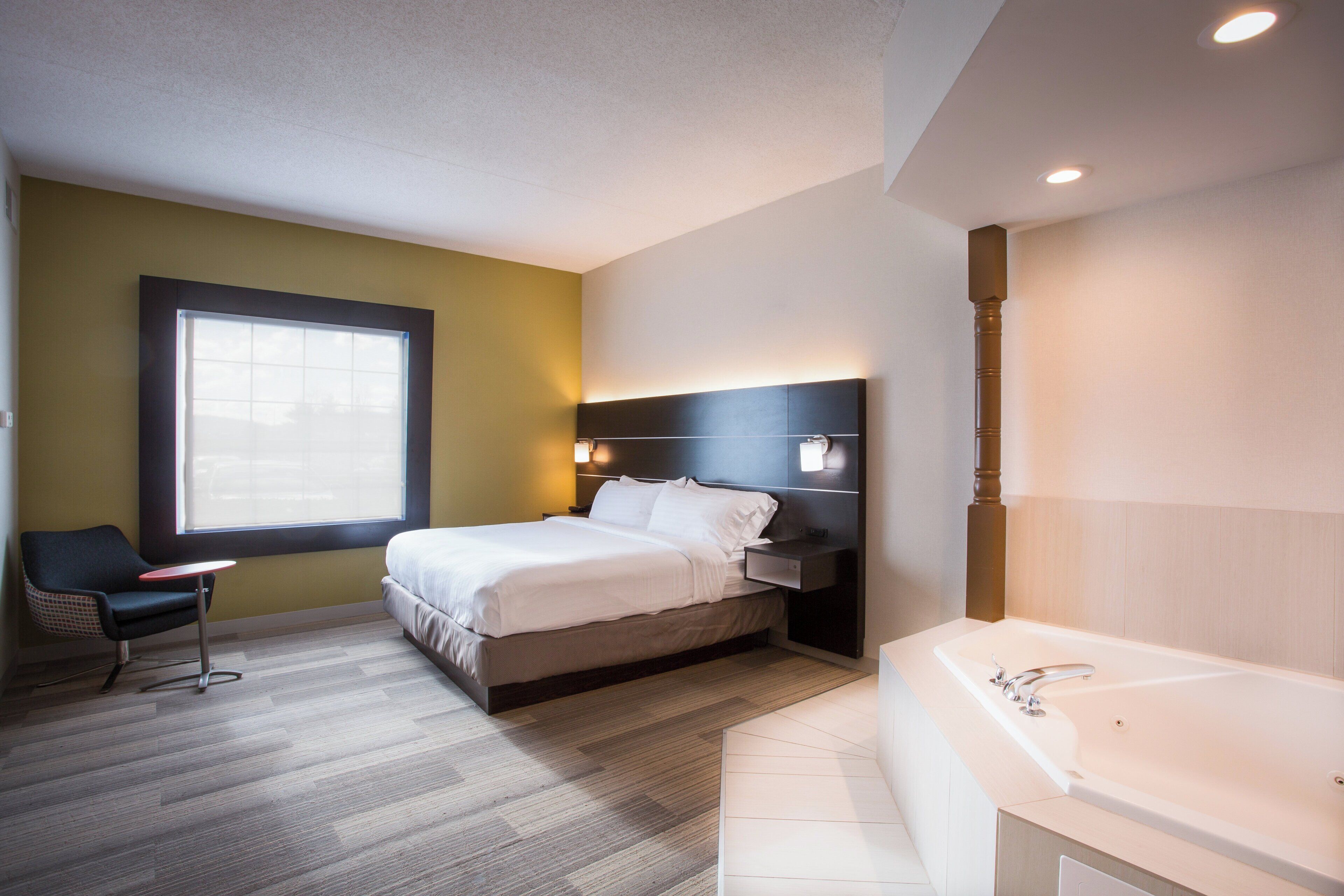 A hotel room with a bed, a chair, a desk, a large window, and a bathtub.
