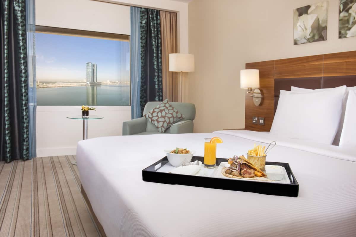 A hotel room with a bed, a dining table with breakfast, a view of a cityscape, and a window with curtains.