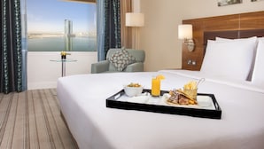 A hotel room with a bed, a dining table with breakfast, a view of a cityscape, and a window with curtains.