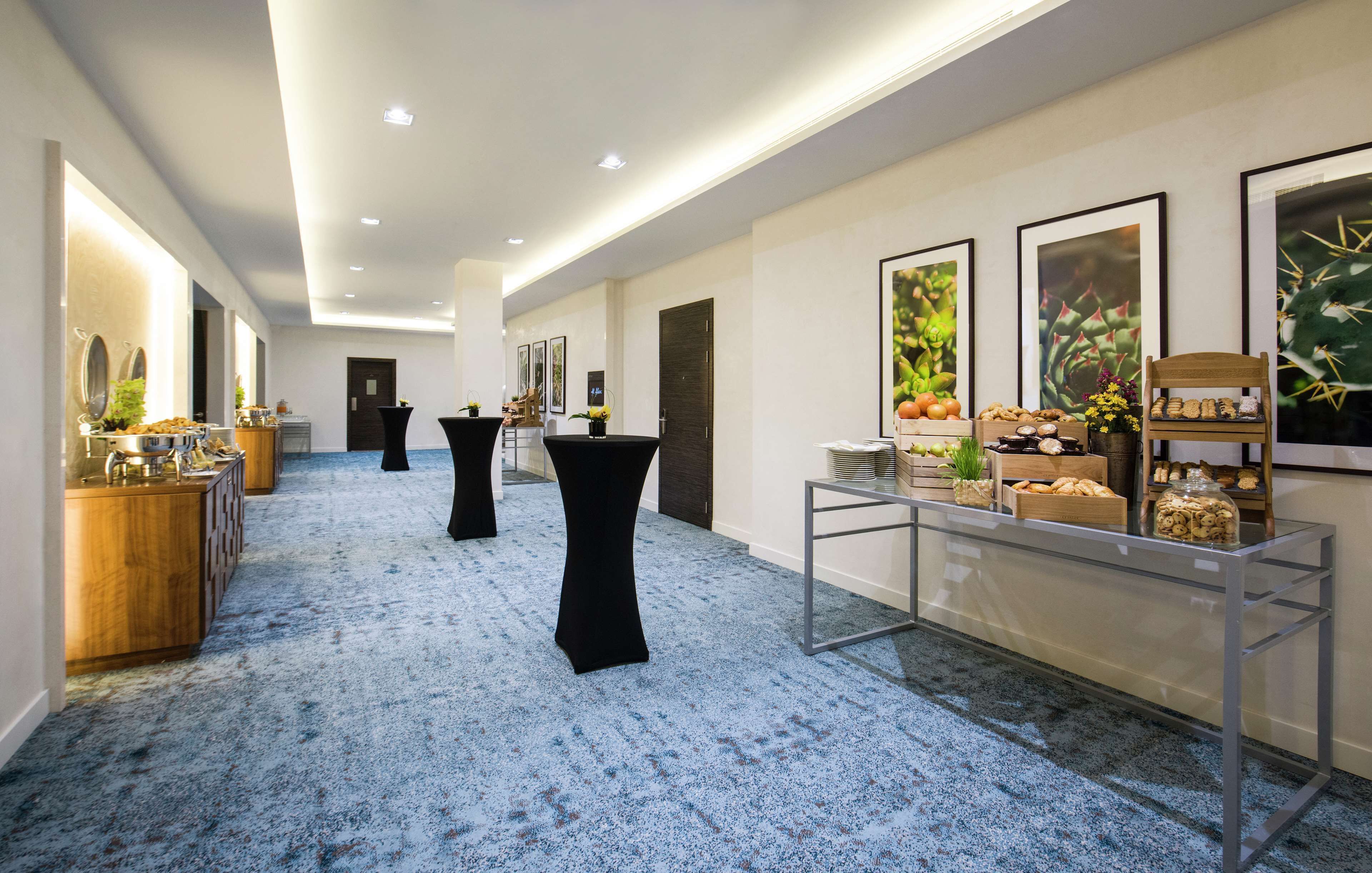 A hallway with a blue carpet, black round tables, and a glass display case with food items.