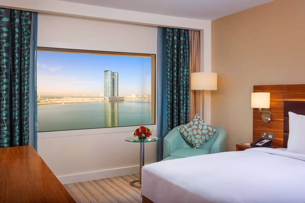 A hotel room with a bed, a small table with a vase of flowers, a chair, a nightstand with a lamp, and a view of a cityscape through a large window.