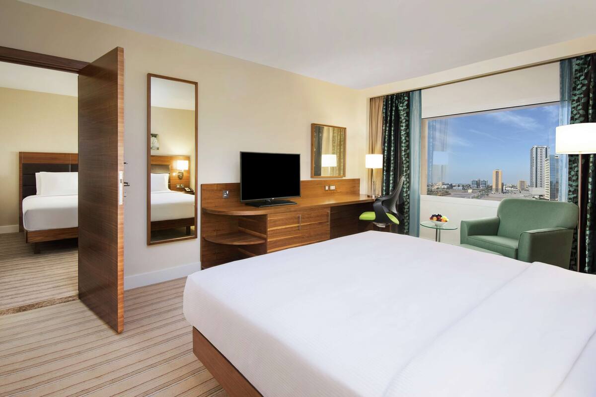 A hotel room with a bed, a desk with a TV, a chair, a small table with a lamp, and a view of a cityscape.