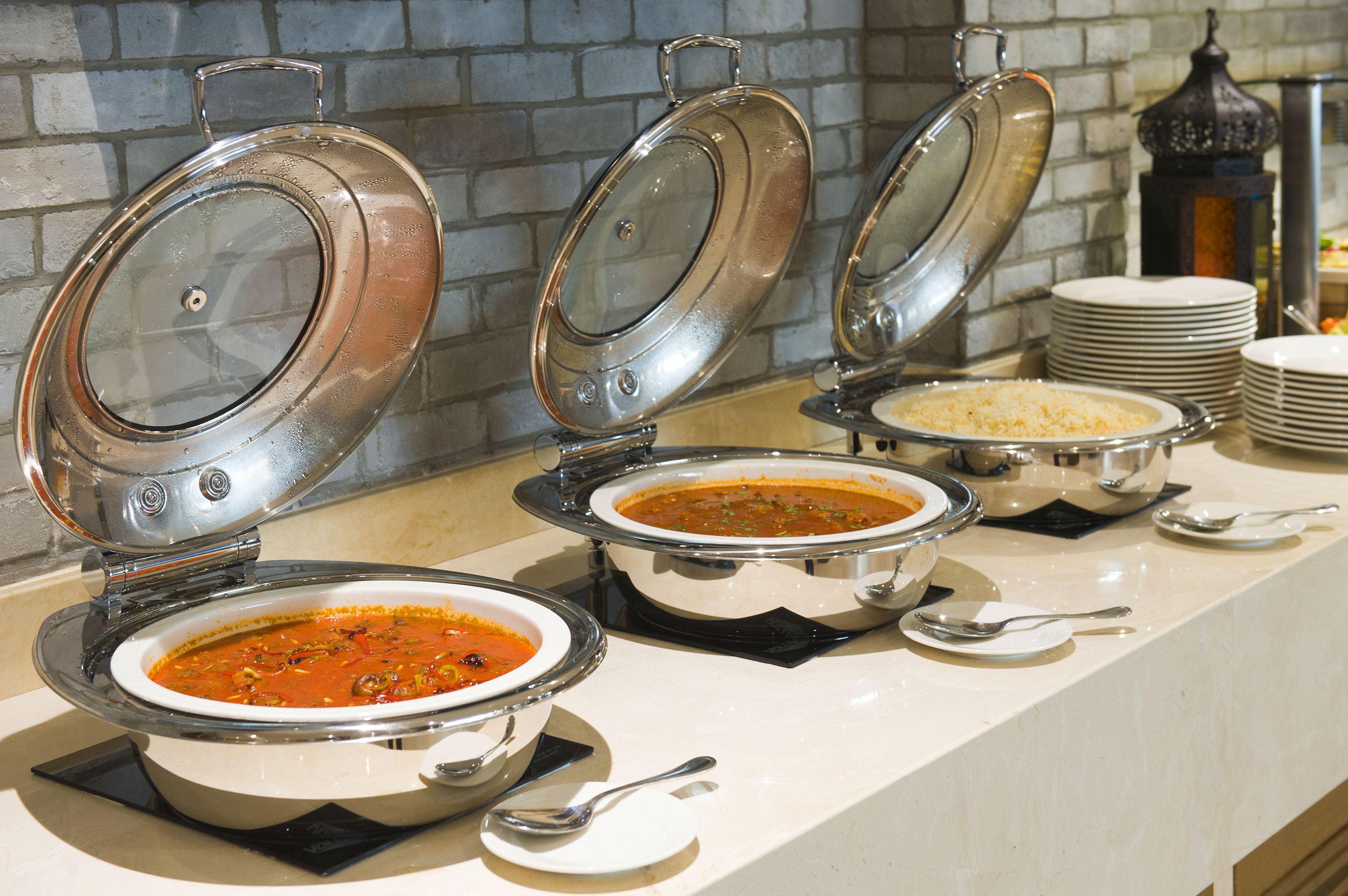 Un buffet proposant divers plats présentés dans des plats de service en métal, avec un mur de briques en arrière-plan et une pile d’assiettes.