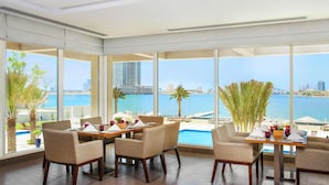 A dining area with a view of the sea and city skyline, featuring large windows, modern furniture, and a poolside setting.
