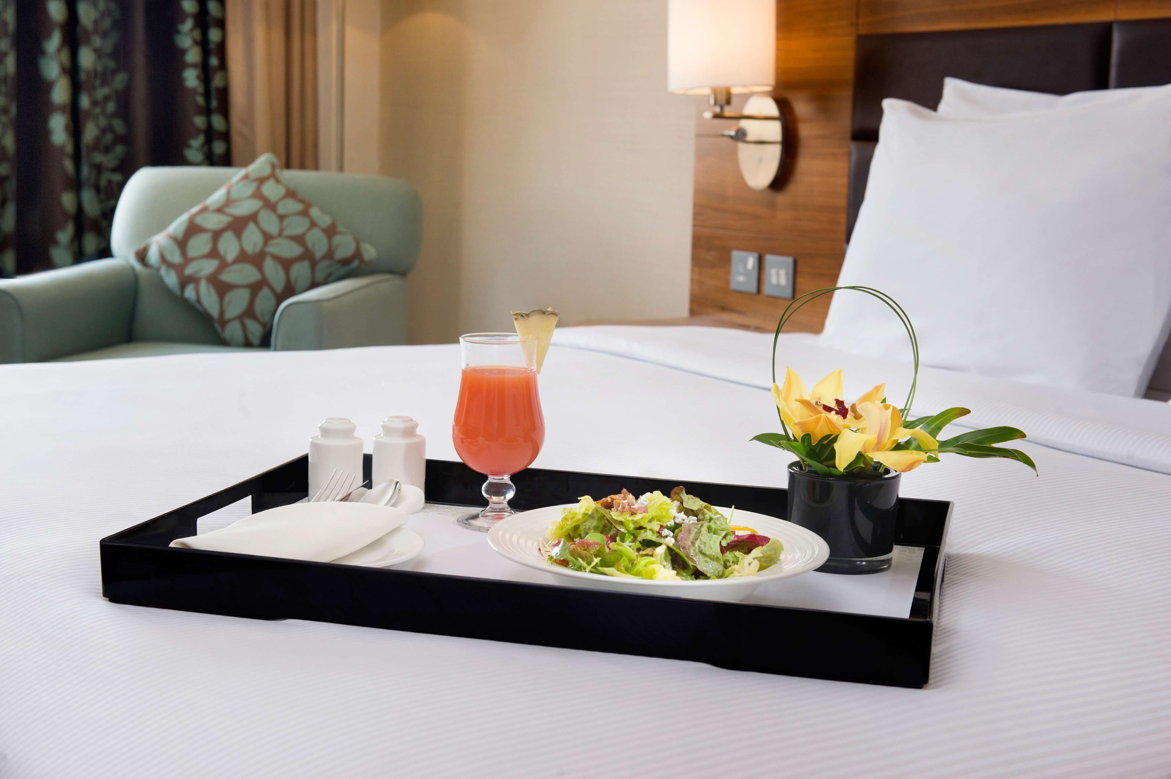 Una bandeja de desayuno ordenada sobre una cama de hotel, con una ensalada, un vaso de jugo y un pequeño jarrón de flores.