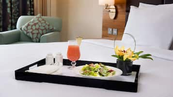 Una bandeja de desayuno ordenada sobre una cama de hotel, con una ensalada, un vaso de jugo y un pequeño jarrón de flores.