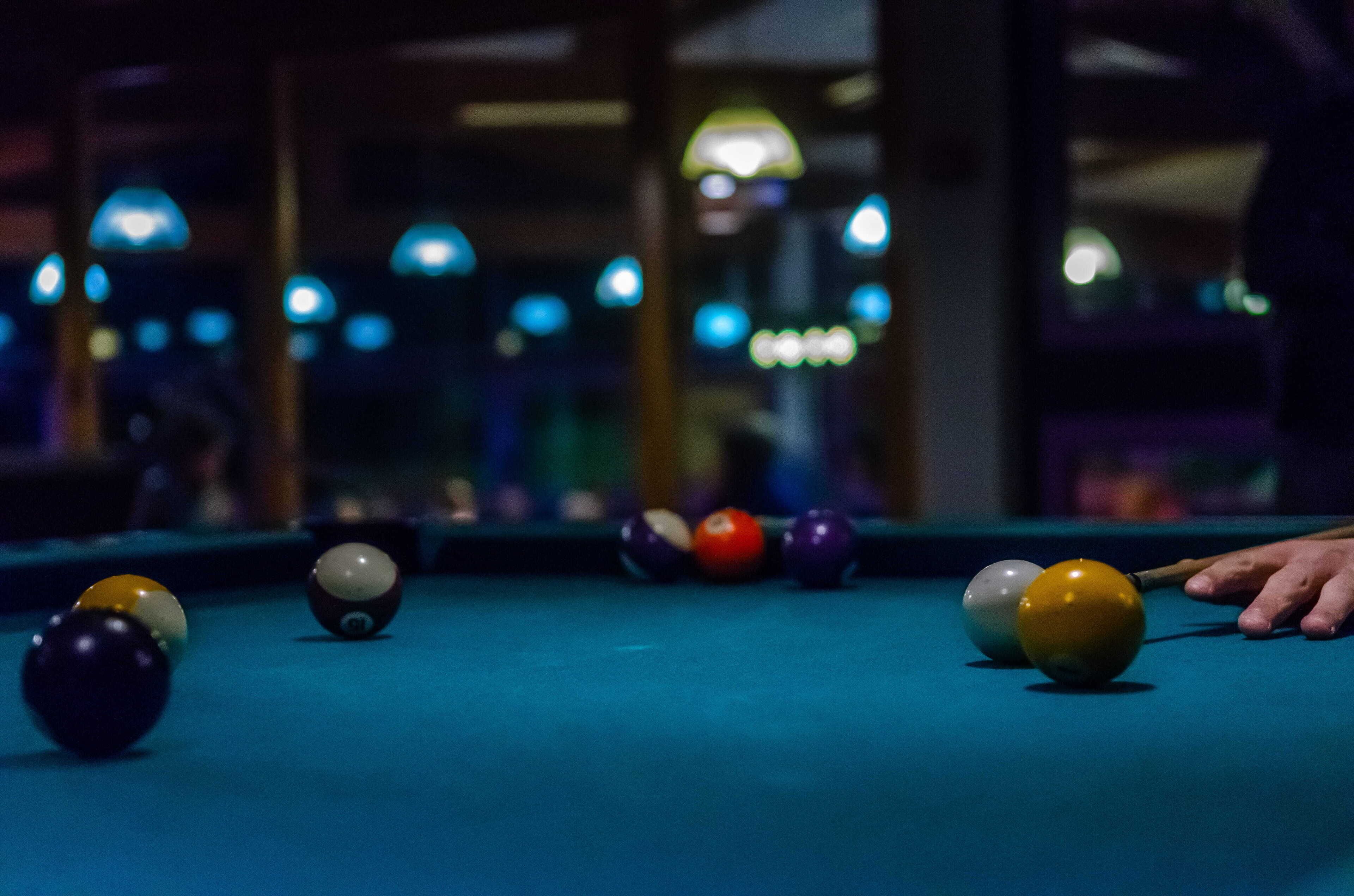 A pool table with balls in various colors, a pool cue, and a person's hand reaching for a ball.