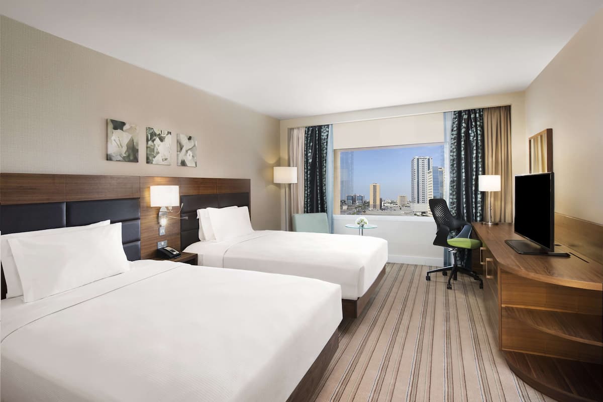 A hotel room with two beds, a desk, a chair, and a large window with a city view.