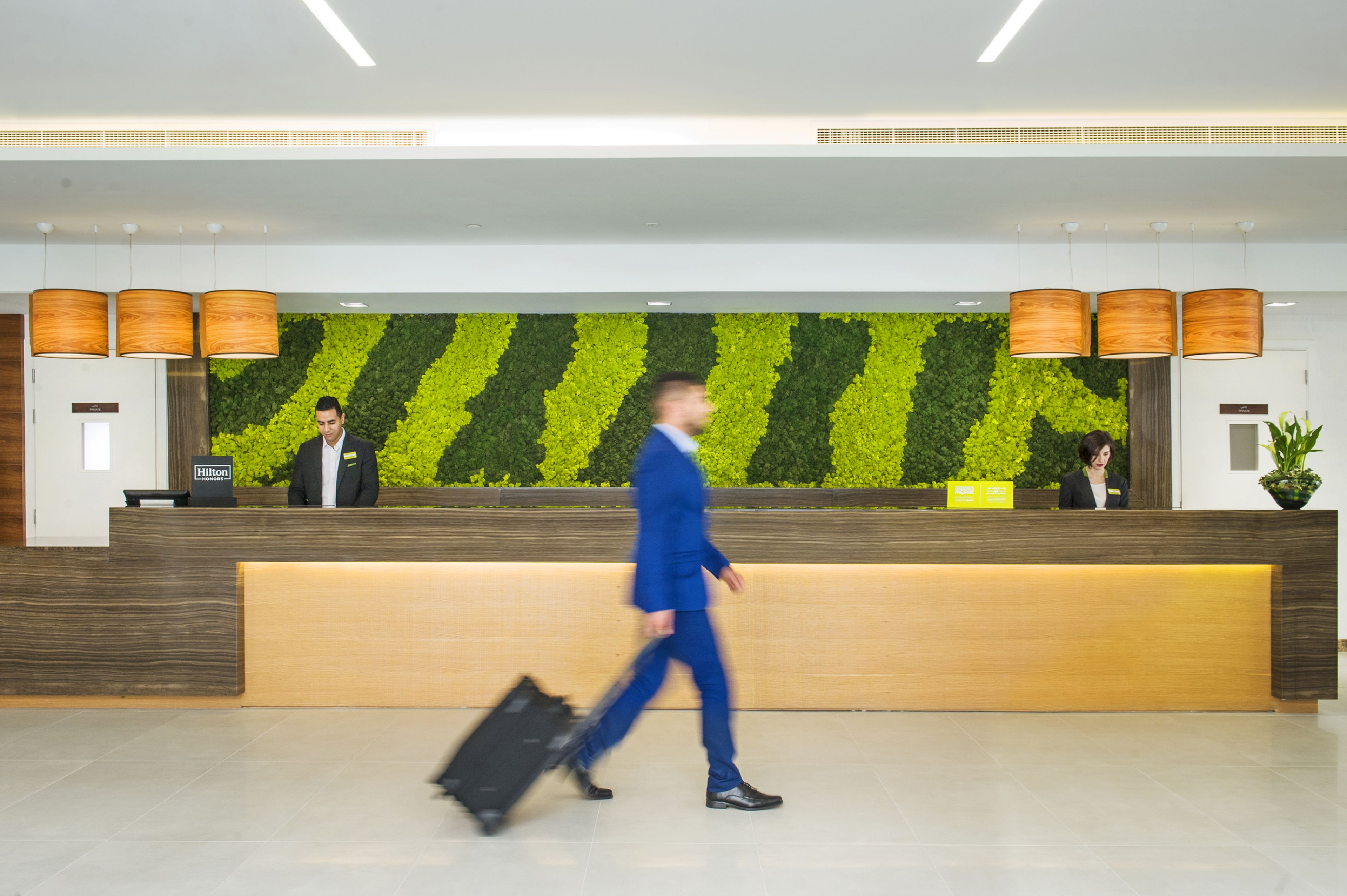 Un hall d’accueil d’hôtel avec un mur végétal, un comptoir d’accueil en bois et deux employés.