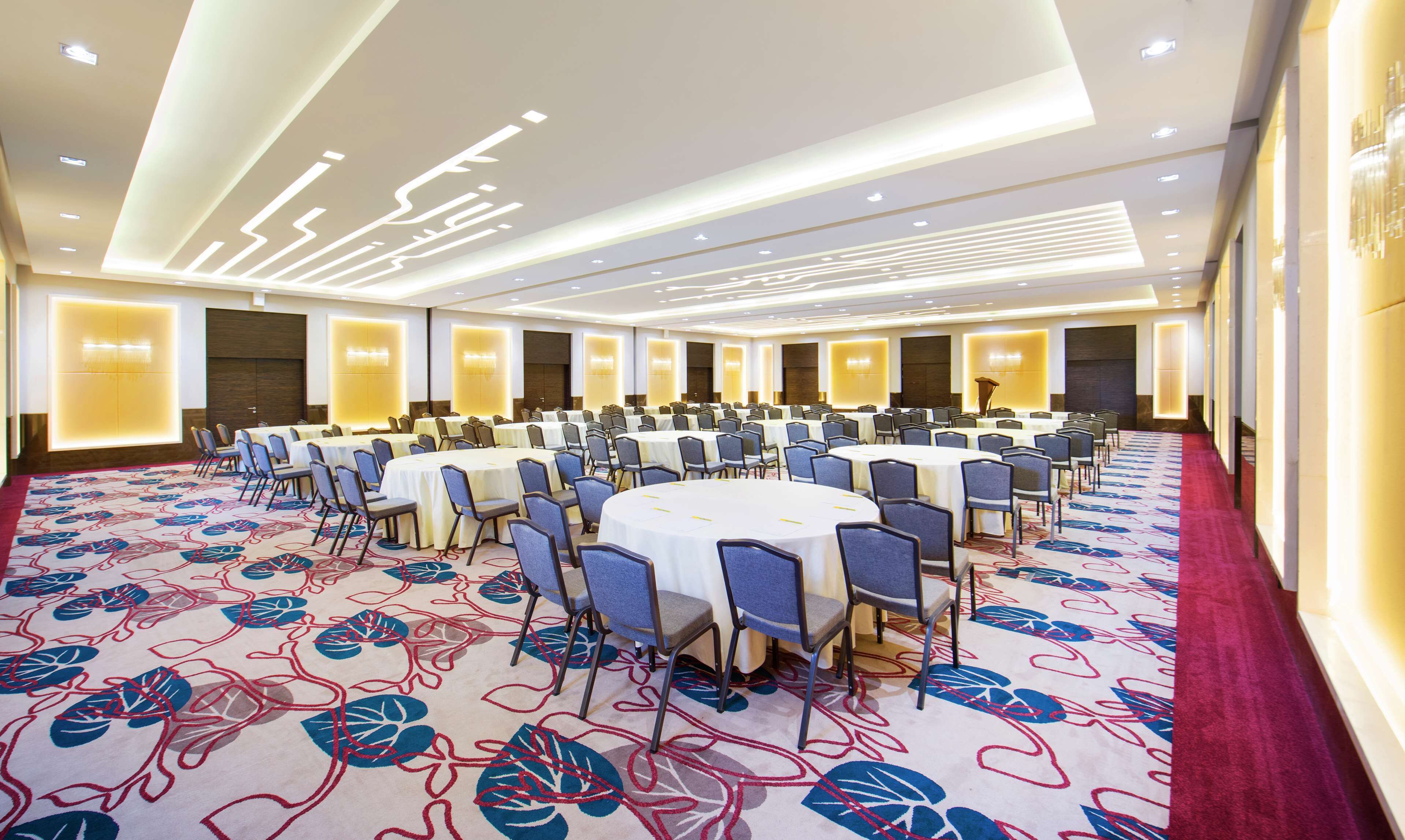 Un salón de conferencias con mesas y sillas redondas, alfombra estampada e iluminación empotrada en el techo.